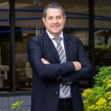 A professional man stands confidently outside an office building, wearing a suit and tie, with his arms crossed. Lush green plants are visible in the foreground, and large windows behind him provide a view of the office environment.