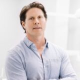 A professional-looking man with medium-length dark hair stands confidently with his arms crossed. He is wearing a light blue shirt and is positioned against a bright, modern background that suggests a clean environment.