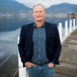 A smiling man stands on a wooden pier beside a calm lake, wearing a dark blazer over a blue shirt and jeans. The background features mountains and a cloudy sky, creating a serene atmosphere.