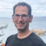 A man with glasses smiles at the camera, standing outdoors near a body of water. He has short dark hair and is wearing a dark shirt. The background features a scenic waterfront view with rocks and trees.