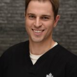 A smiling male dentist wearing a black dental uniform with a logo, standing against a textured gray background.