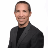 A man with short hair smiles confidently while wearing a dark blazer over a black shirt. The background is plain white, emphasizing his professional appearance.