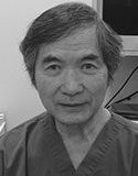 A portrait of an older Asian male wearing medical scrubs, looking directly at the camera with a neutral expression. The background is indistinct, focusing on the individual.