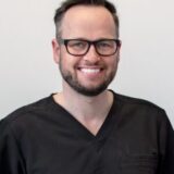 A smiling man wearing glasses and a black scrub top stands against a light background.