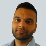 A man with a short beard and dark hair gazes slightly to the side, wearing a light blue shirt against a pale background.