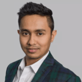 A professional portrait of a man with short black hair, wearing a dark plaid blazer over a white shirt, smiling confidently. The background is a neutral gray, focusing attention on his face and attire.