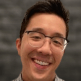 A smiling person with short hair and glasses is wearing a checkered shirt, posed against a blurred dark background.