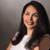 A woman with long dark hair smiles at the camera. She is wearing a white top and has a neutral background.