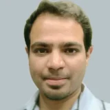 A close-up portrait of a man with dark hair and a slight smile, wearing a light blue shirt against a plain gray background.