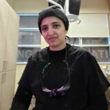 A healthcare professional wearing a black uniform and a head covering stands in a clinical setting. She has specialized glasses around her neck and appears focused, suggesting an environment related to dentistry or a similar field.