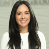 A smiling woman with long black hair and wearing a white sweater poses in front of a softly blurred background.