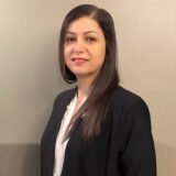 A woman with long dark hair wearing a black blazer and a white top stands against a neutral wall, smiling confidently.
