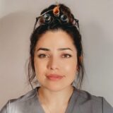 A woman with dark hair tied back is wearing glasses on her head and a grey medical scrub top. She has a neutral expression, standing against a light background.