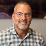 A smiling man with a beard and glasses, wearing a checked shirt, is seated in an office setting, with an abstract background.