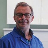 A middle-aged man with glasses and gray hair smiles confidently while wearing a blue lab coat, standing in a well-lit room with cabinets in the background.