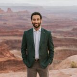 A man stands confidently in front of a scenic canyon landscape, wearing a dark blazer over a light shirt and grey pants. The vast, rugged terrain stretches behind him, showcasing layers of red rock formations and a dramatic sky.