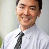 A smiling man with short dark hair is wearing a light blue dress shirt and a patterned tie. He is standing outdoors, with a soft-focus background suggesting an office or urban environment.