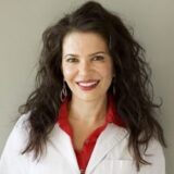 A smiling woman with long, wavy dark hair is wearing a white lab coat over a red blouse. She has dangling earrings and stands in front of a neutral background.