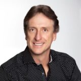 A person with medium-length brown hair smiles at the camera, wearing a patterned black shirt. The background is a light gradient, enhancing the focus on the individual's friendly expression.