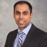 A man in a dark suit and light blue shirt smiles confidently against a textured gray background. His hair is neatly styled, and he wears a striped tie, presenting a professional appearance.