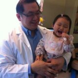 A smiling adult wearing a lab coat holds a cheerful baby, who is laughing with a pacifier in her mouth. The setting appears cozy, with playful items in the background.