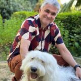 A smiling man in a colorful shirt is sitting outdoors beside a fluffy white dog. They are surrounded by greenery, and the man is gently petting the dog.