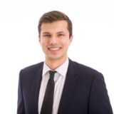 A young man in a dark suit and tie smiles confidently against a light background. He has short, dark hair and is standing in a professional pose.