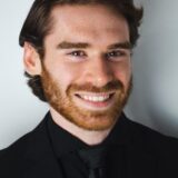 A young man with a reddish beard and light brown hair smiles confidently while dressed in a black suit and tie against a neutral background.