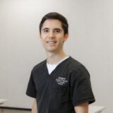 A smiling male healthcare professional wearing dark scrubs stands against a neutral background.