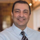 A smiling man wearing a light blue dress shirt and a striped tie poses for the camera. The background features warm lighting and a blurred interior setting.