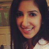 A smiling woman with long dark hair is sitting indoors, wearing a white shirt. The background shows a blurred interior space, suggesting a social or dining setting.