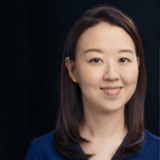 A woman with shoulder-length dark hair smiles at the camera, wearing a blue top. The background is dark, highlighting her friendly expression.