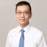 A man wearing glasses, a white shirt, and a blue tie smiles at the camera against a neutral background.