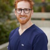 A smiling man with red hair and glasses wears dark blue scrubs, standing outdoors against a blurred background of greenery.