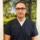 A man in dark scrubs stands in front of a backdrop featuring a sunny, green landscape with trees. He has short, dark hair and is wearing glasses, looking directly at the camera with a serious expression.