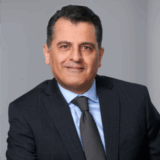 A professional man in a dark suit and light blue shirt poses confidently against a neutral gray background. He has dark hair, a slight smile, and is wearing a tie, suggesting a business or corporate setting.