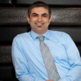 A smiling man with short dark hair wearing a light blue dress shirt and a patterned tie sits casually against a dark background.