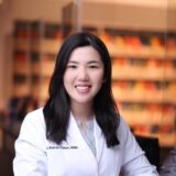 A woman with long, dark hair smiles while wearing a white lab coat. She stands in a dental office with colorful shelves in the background, suggesting a professional and welcoming environment.