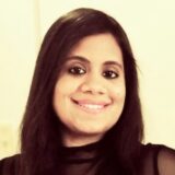 A smiling woman with long black hair poses for a photo, wearing a black outfit. The background is softly blurred, focusing on her cheerful expression.