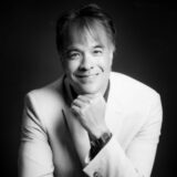 A smiling man in a light-colored suit rests his chin on his hand, exemplifying a thoughtful pose against a dark background. The image is in black and white, highlighting his features and expression.