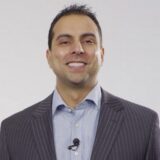 A smiling man wearing a striped suit and a collared shirt stands against a neutral background. He appears confident and approachable, with a microphone attached to his shirt.