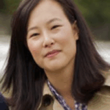 A woman with long, dark hair smiles softly while wearing a beige coat over a checkered shirt. She is outdoors with a blurred natural background.