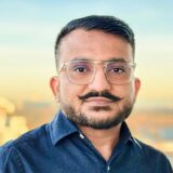 A man with glasses and a neatly styled mustache smiles against a blurred outdoor background. He wears a blue shirt and has short hair, conveying a friendly and professional demeanor.