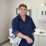 A smiling dental professional stands in a modern dental office, leaning on an examination chair. The background features dental equipment and a clean, organized workspace, conveying a friendly and professional atmosphere.