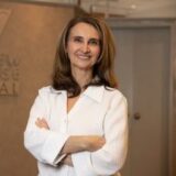 A smiling woman with long hair stands confidently with her arms crossed, wearing a white blouse. She is positioned in an office environment, with soft lighting and a neutral backdrop.