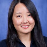 A smiling young woman with long dark hair, wearing a black shirt with a gold necklace, poses against a blue background.