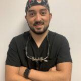 A smiling young man wearing a black medical scrub and a patterned surgical cap stands with his arms crossed. He has dental glasses resting around his neck and is against a plain background.