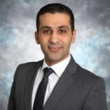 A professional man wearing a gray suit and tie poses for a headshot, smiling confidently against a blurred blue and gray background.