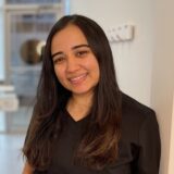 A woman with long, dark hair smiles while standing in a well-lit indoor space. She is wearing a black top and appears approachable and friendly.