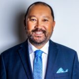 A professionally dressed man with a beard smiles confidently at the camera. He wears a dark suit, a crisp white shirt, and a blue tie with a matching pocket square. The background is a light color, emphasizing his friendly demeanor.